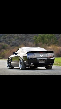 the rear end of a black and white mustang on a road with trees in the background