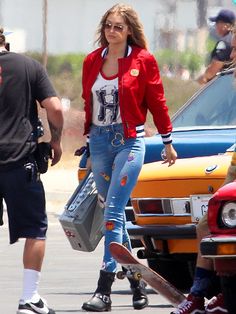 a woman in red jacket and jeans walking down the street with her skateboard behind her