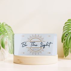 a sign that says be the light on it next to some plants and a potted plant