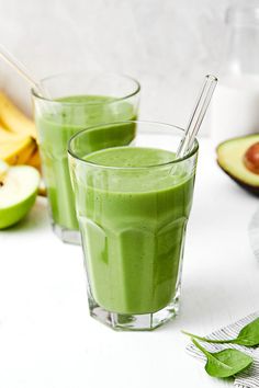 two glasses filled with green smoothie next to sliced bananas and avocado slices