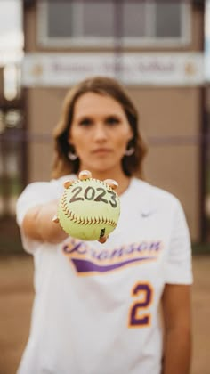 a woman holding a softball in her right hand and wearing a white shirt with the number two on it