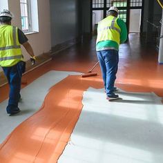 two men in yellow vests and blue jeans are painting the floor with orange paint