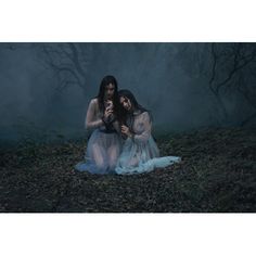 two women sitting on the ground in front of some dark trees and foggy sky