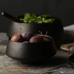 some plums are in a black bowl on a table next to other fruits and vegetables