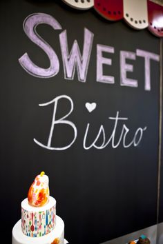 a white cake sitting on top of a table next to a chalkboard with the words sweet bistro written on it