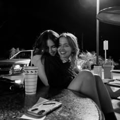 two women sitting at a table with their arms around each other, smiling and hugging