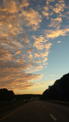 the sun is setting behind some trees on the side of the road with clouds in the sky