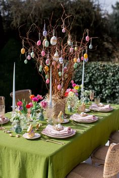 the table is set for easter dinner with green linens and colorful eggs hanging from branches