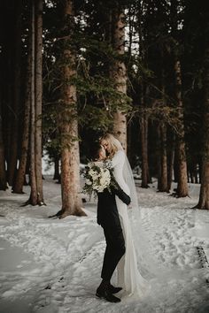 a bride and groom standing in the snow with trees behind them, looking at each other