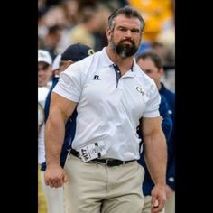 a man with a beard wearing a white shirt and khaki pants walks on the sidelines