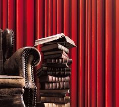 a chair sitting next to a stack of books on top of a wooden table in front of a red curtain