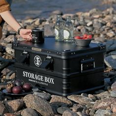 a black storage box sitting on top of rocks next to a body of water with two bottles