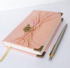 a pink notebook with a gold bee on it next to a pen and paper clip