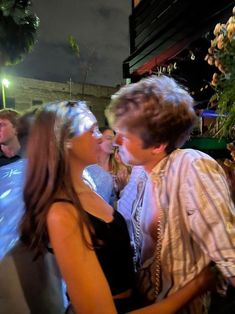 a man and woman kissing each other in front of a crowd at night time with lights on