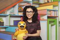 a woman holding a yellow stuffed animal in front of colorful bookshelves and stairs