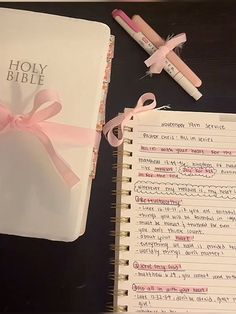 an open bible with pink ribbon and two pencils next to it on a table