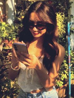 a young woman wearing sunglasses is looking at her cell phone in front of some plants