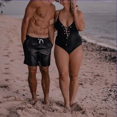 a man and woman in bathing suits walking on the beach with one holding his nose