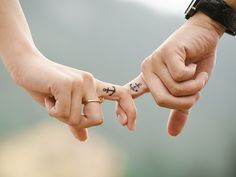 two people holding hands with small tattoos on their thumbnails, one has an anchor and the other is a cross