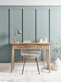 a desk with a chair, lamp and vase on it in front of a blue paneled wall