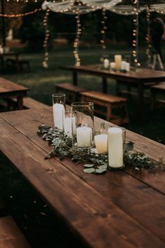 candles and greenery sit on an outdoor wooden table in front of string - lights