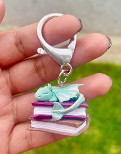 a hand holding a tiny book charm with a lizard on it's back, sitting on top of a stack of books