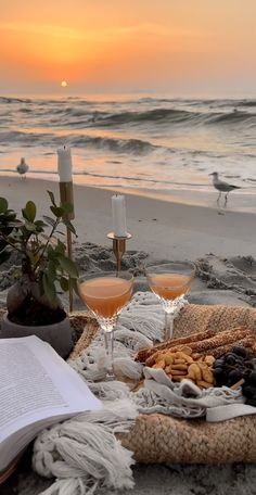 two glasses of wine are sitting on a blanket at the beach with an open book