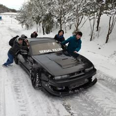 four people standing next to a black car on snow covered road with trees in the background