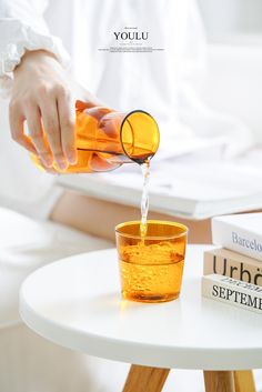 a person pouring orange juice into a glass on top of a white table next to a stack of books