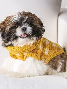 a small dog wearing a yellow sweater sitting on a white couch with its tongue out