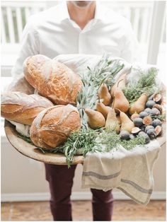 a person holding a platter full of bread, fruit and veggies in front of a window