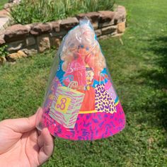 a hand holding up a plastic cone with barbie dolls on it in the middle of a yard