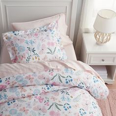 a bed with pink and blue flowers on it next to a white night stand holding a lamp