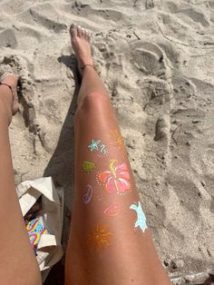 a woman's legs with colorful tattoos on them sitting in the sand at the beach