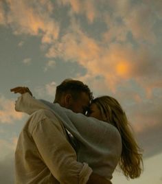 a man and woman are hugging under the sky with clouds in the background at sunset