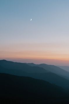 the sun is setting in the distance over some hills and mountains with a half moon