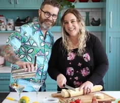 a man and woman standing in front of a cutting board with cheese on it, smiling at the camera