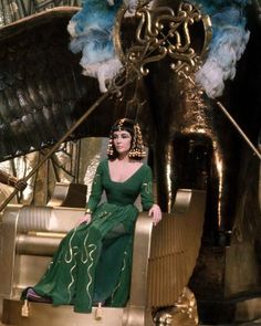 a woman in a green dress sitting on top of a gold car with feathers hanging from it's wings