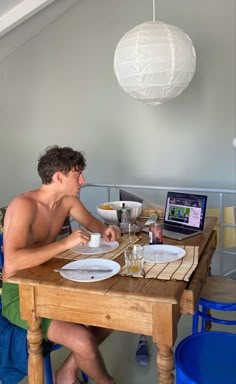 a shirtless man sitting at a wooden table with plates and cups in front of him