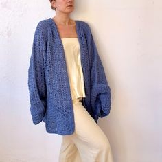 a woman leaning against a white wall wearing a blue cardigan sweater and cream pants