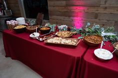 a red table topped with lots of food and desserts next to a wall covered in pink lights