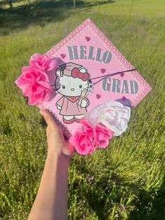 someone holding up a hello kitty graduation cap with pink flowers in front of green grass