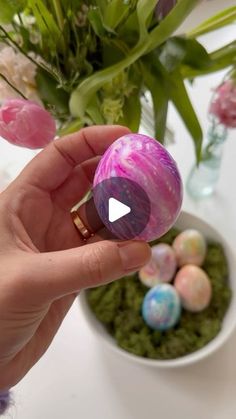 a person holding an easter egg in front of some flowers and eggs on a table