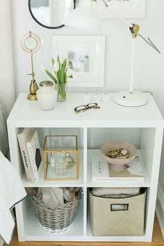 a white shelf with baskets, books and other items on it next to a lamp