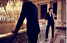 a woman standing next to a man in a suit and tie on a balcony with chandelier