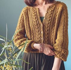 a woman standing next to a plant wearing a brown cardigan sweater and pleated skirt