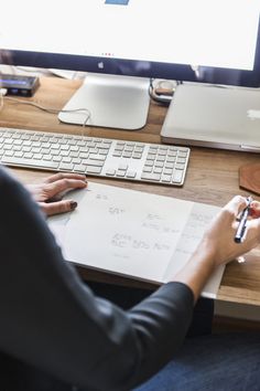 a person sitting at a desk writing on a piece of paper next to a computer