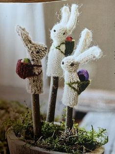 three knitted rabbits are sitting on top of sticks in a pot with moss and flowers