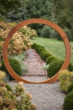 a circular wooden sculpture in the middle of a gravel path surrounded by bushes and trees