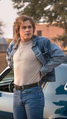 a young man standing in front of a blue car wearing jeans and a denim jacket